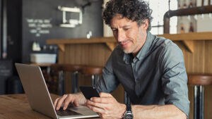 A man sits in a coffee shop using his laptop and cellphone to access personal information. 