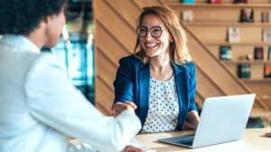 Male and female sitting at a table with laptop, shaking hands.