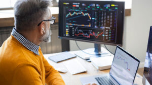 A man in his 50s wearing glasses sits at his desk reviewing stock market information on a large monitor screen and his laptop and thinks about strategies for surviving market turbulence.