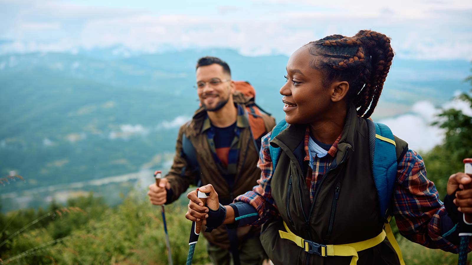 Young man and woman hiking with walking sticks at the top of a mountain smiling.